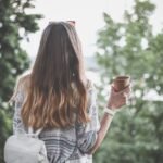 Back view of a young woman with long hair holding a coffee cup outdoors in a tranquil park.